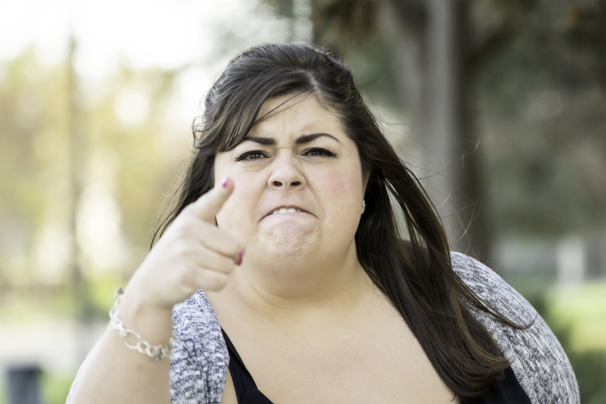 Angry obese woman directly looking towards camera, pointing her finger, a blurred background of outside on a sunny day