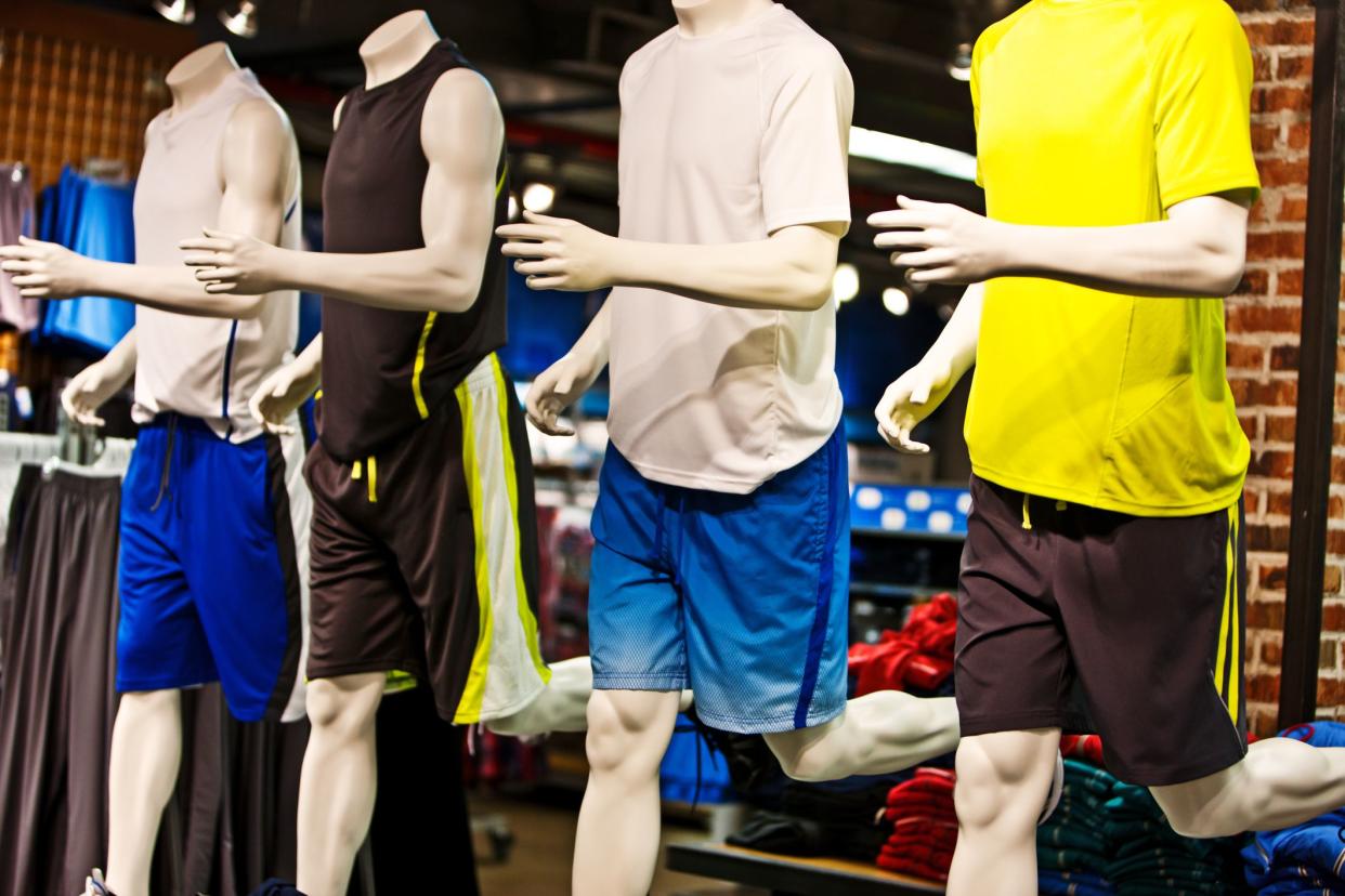 Male exercise clothing on four mannequins in a trendy store with clothing racks and piles of clothes on display in the background