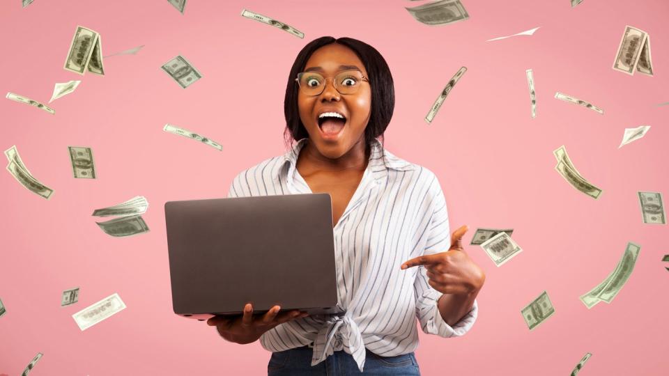 A person with mouth open and eyes wide, holding a laptop as paper money falls around them. 