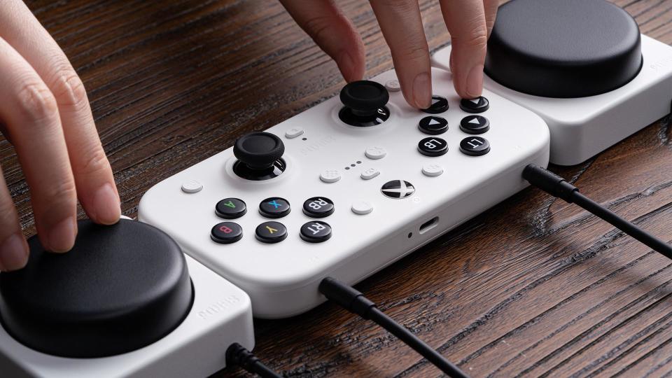 A person's hands controlling the 8BitDo Lite SE 2.4G Wireless Controller, which has all its buttons on the top surface. Two Super Buttons flank it on either side.