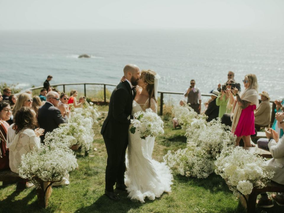A bride and groom kiss in front of the ocean as their guests cheer.