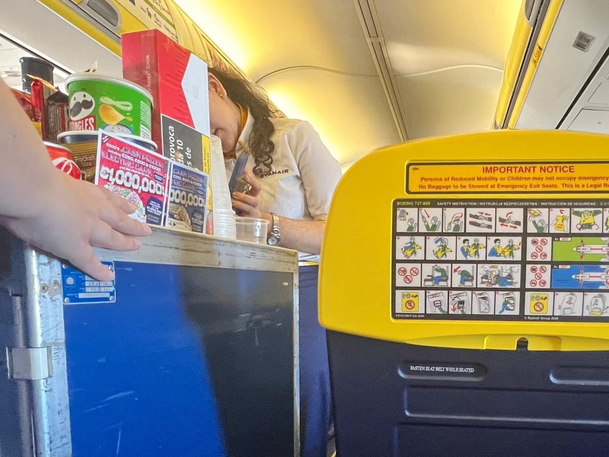 Two Ryanair flight attendants push a trolley down the aisle, with pringles and scratch cards and cigarettes on top, from the aisle seat's point of view.