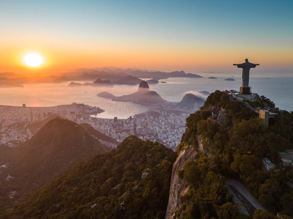 Aerial view of Rio de Janeiro showcasing the Christ the Redeemer statue on top of Corcovado Mountain, with Sugarloaf Mountain on the horizon during sunrise.