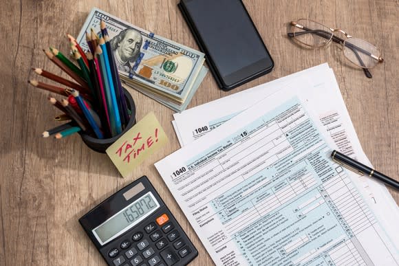 Tax forms, calculator, money, pencils, and glasses on a table.