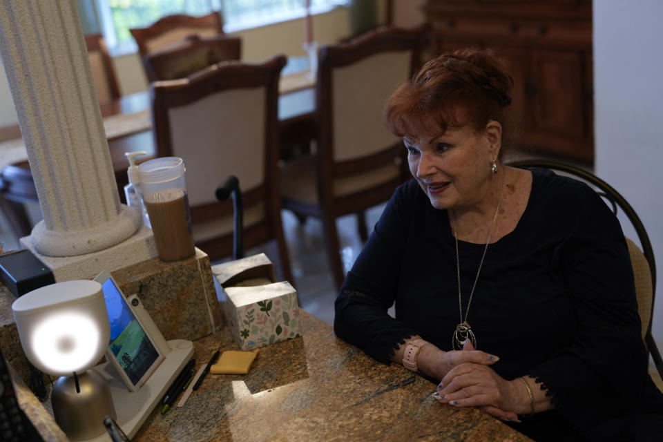 Deanna Dezern, 83, prompts her ElliQ, left, a tabletop device that uses artificial intelligence to conduct human-like conversations, to speak to visiting journalists, inside her home in Tamarac, Fla., Thursday, Dec. 7, 2023. Dezern is among the first in the country to receive the robot ElliQ, whose creators, Intuition Robotics, and senior assistance officials say is the only device using artificial intelligence specifically designed to lessen the loneliness and isolation experienced by many older Americans. (AP Photo/Rebecca Blackwell)