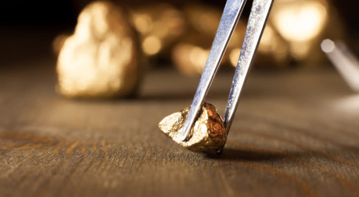 A photo of a gold nugget on a table, being picked up by tweezers, with more gold behind it.