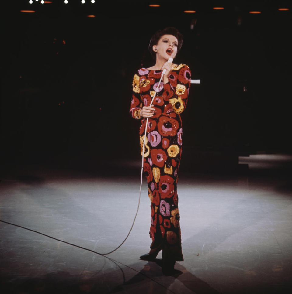 Judy Garland performs on stage, wearing a floral-patterned dress, holding a microphone