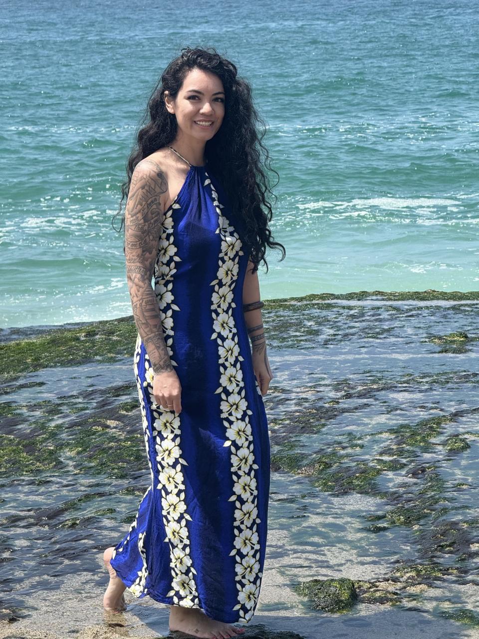 A woman with long, curly hair and tattoos on her arms stands on a rocky beach by the sea wearing a floral dress