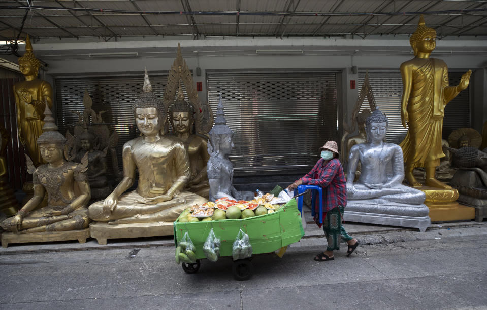 A fruit vendor wearing a protective mask to help curb the spread of the coronavirus pushes her cart past giant Buddhist statues for sale in Bangkok, Thailand, Thursday, May 27, 2021. (AP Photo/Sakchai Lalit)