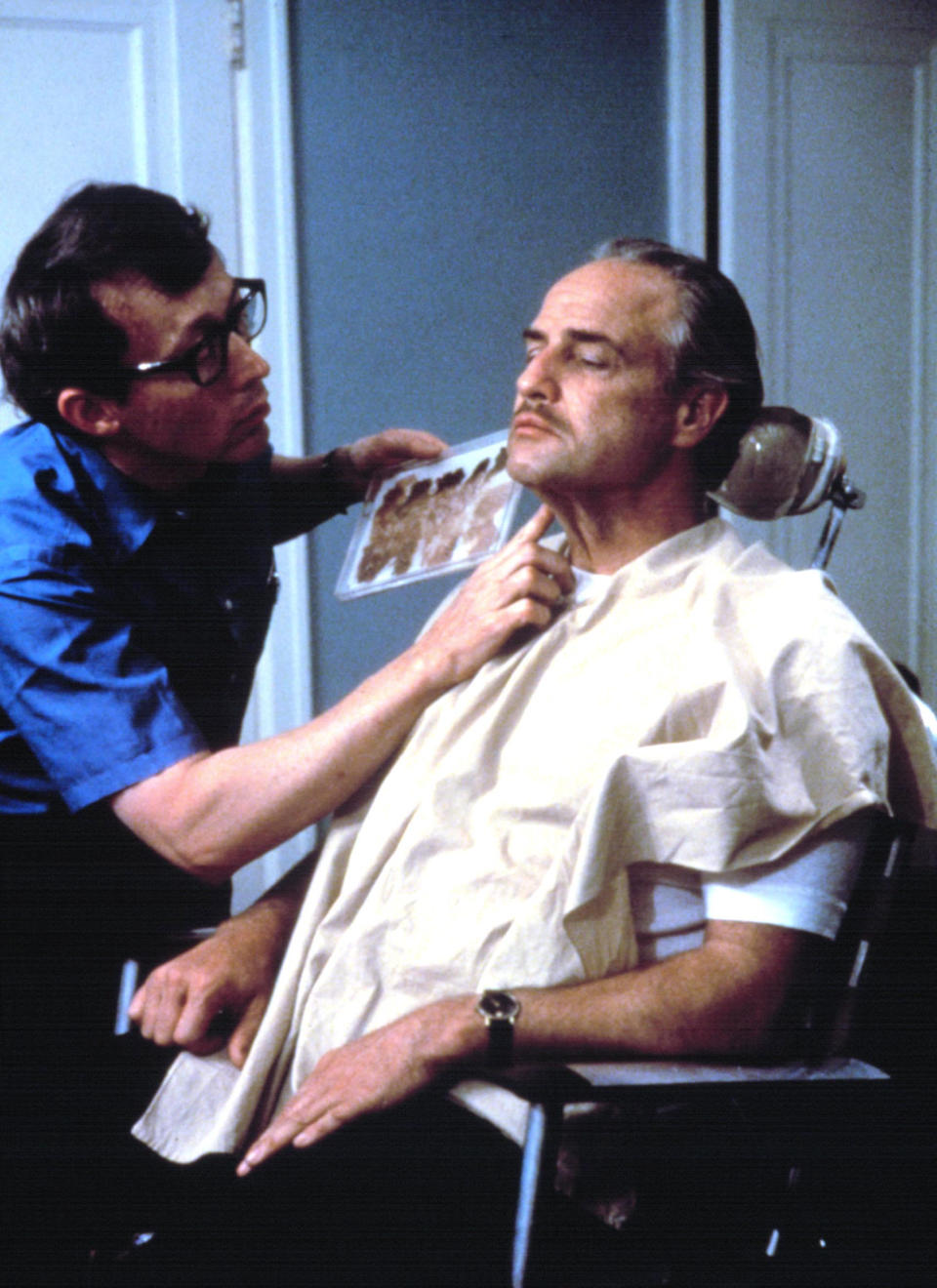 A makeup artist is applying makeup to Marlon Brando, who is seated and wearing a drape over his clothing