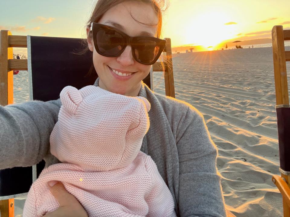 A woman wearing sunglasses takes a selfie at sunset holding her baby.