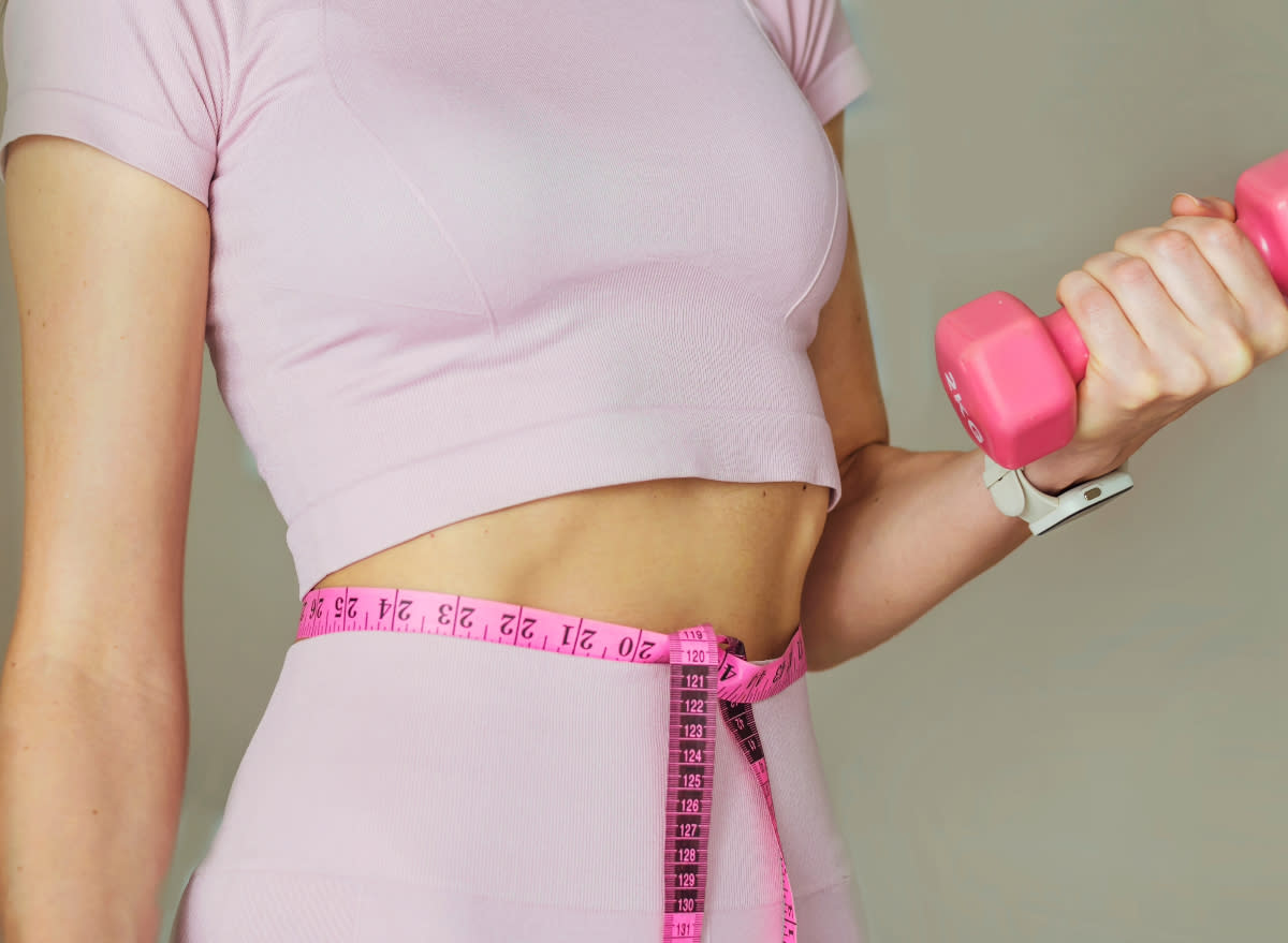 fit woman holding dumbbell with measuring tape around waist, concept of ways to get a leaner body