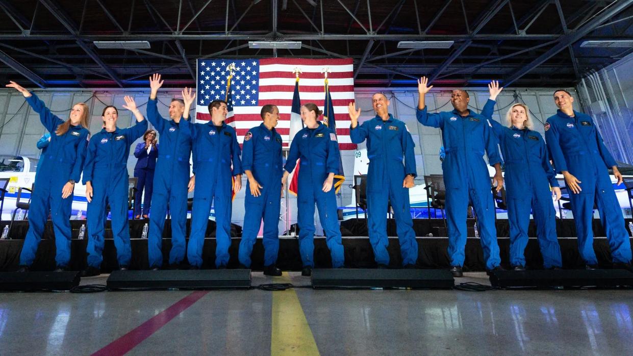  10 astronaut candidates stand in a row in a hangar, in front of an american flag and t-38 jets. 