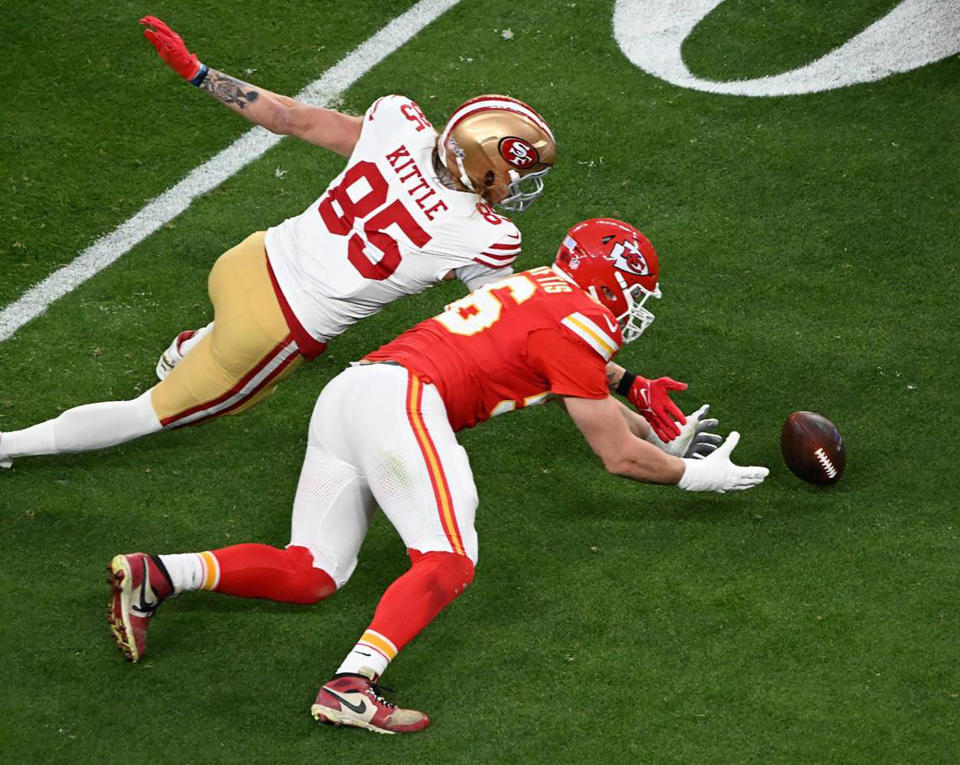 Two football players in action during a game, one dives towards the other who reaches for a football