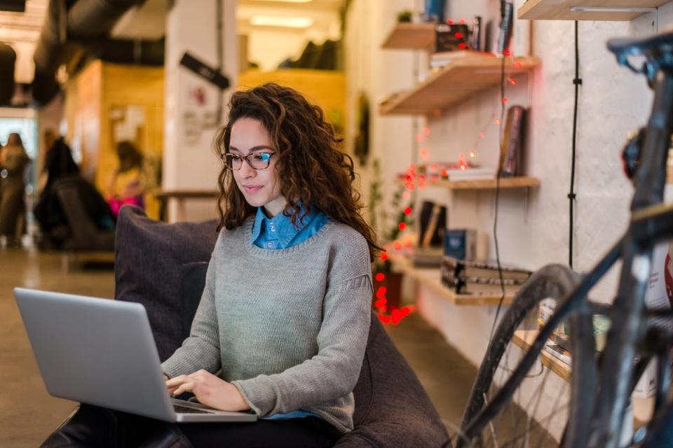 A woman sits on a plush chair in an office typing on a laptop