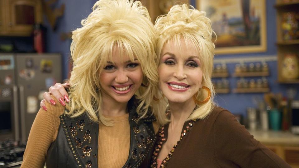 miley cyrus and dolly parton smile for a photo together and hug, cyrus wears a blonde wig styled to look like parton's hair