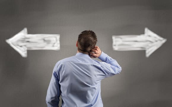 A man in a blue button-down shirt, with his back turned, looking at white arrows on a gray wall pointing left and right