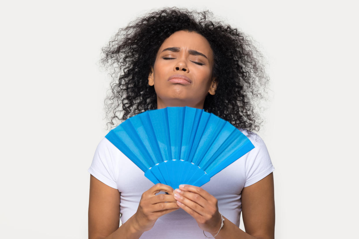 Overheated sweaty african woman holding blue handheld fan posing isolated on grey background. Female feels unwell cooling in summer weather, girl suffers from heat waving fan relief hot day concept