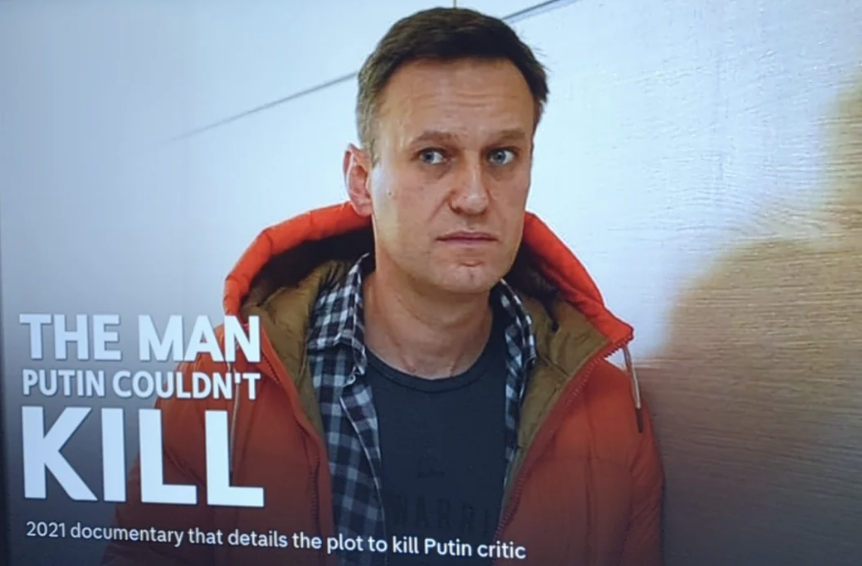 A man stands in front of a documentary title "The Man Putin Couldn't Kill" summarizing a plot against Putin critic