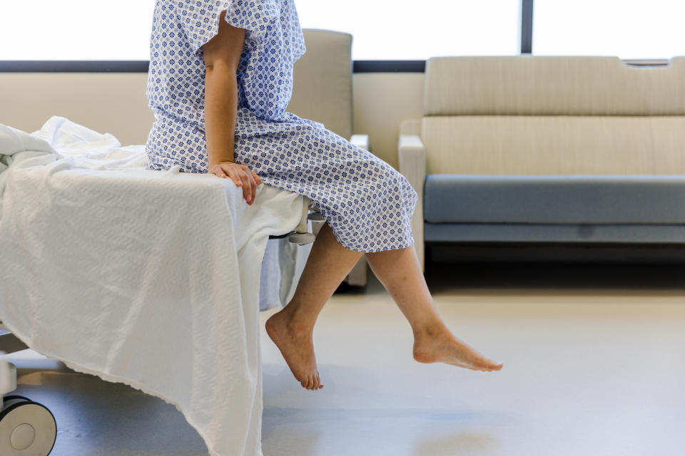 A person, in a patterned hospital gown, is sitting on the edge of a hospital bed with their bare feet hanging off. A sofa is in the background