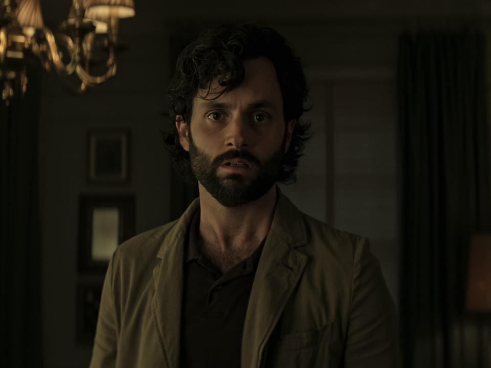 Penn Badgley looking distressed in a dimly lit room with a chandelier in the background on "You"