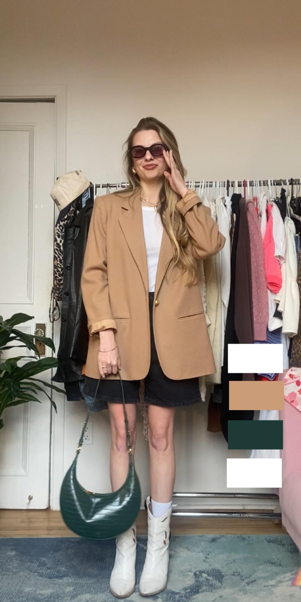 Woman in oversized blazer, mini skirt, and boots, holding purse, standing in a room with clothes rack