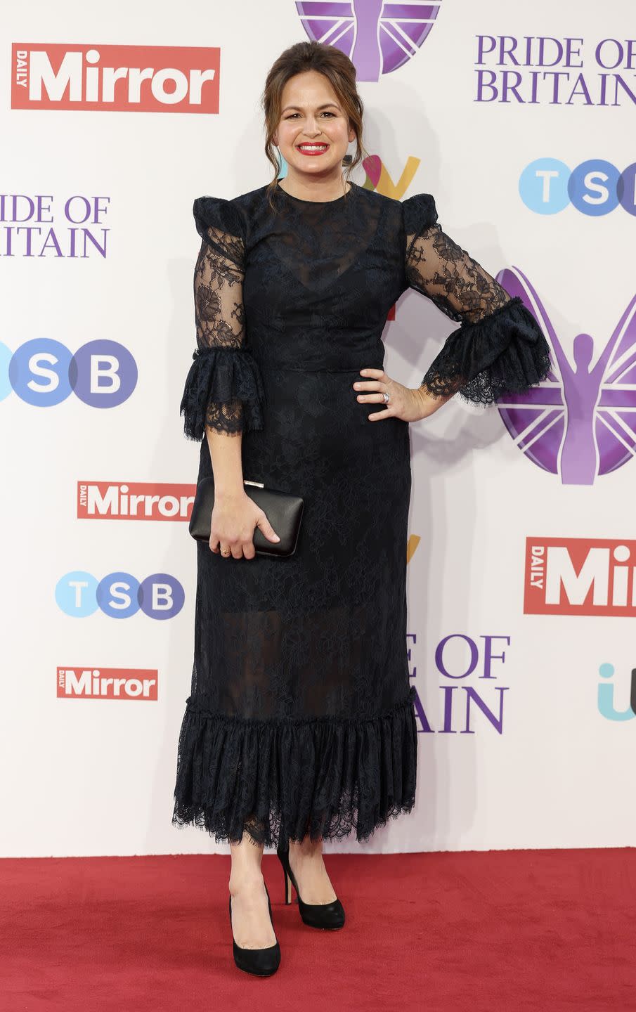 giovanna fletcher, a woman with brown hair in an updo, wearing red lipstick and a black dress, posing with her hand on her hip and smiling at the camera on the red carpet