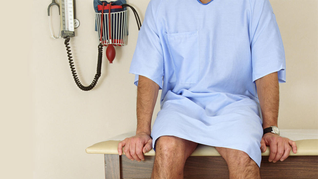 A begowned male patient waits in a doctor's examination room near a blood pressure gauge.