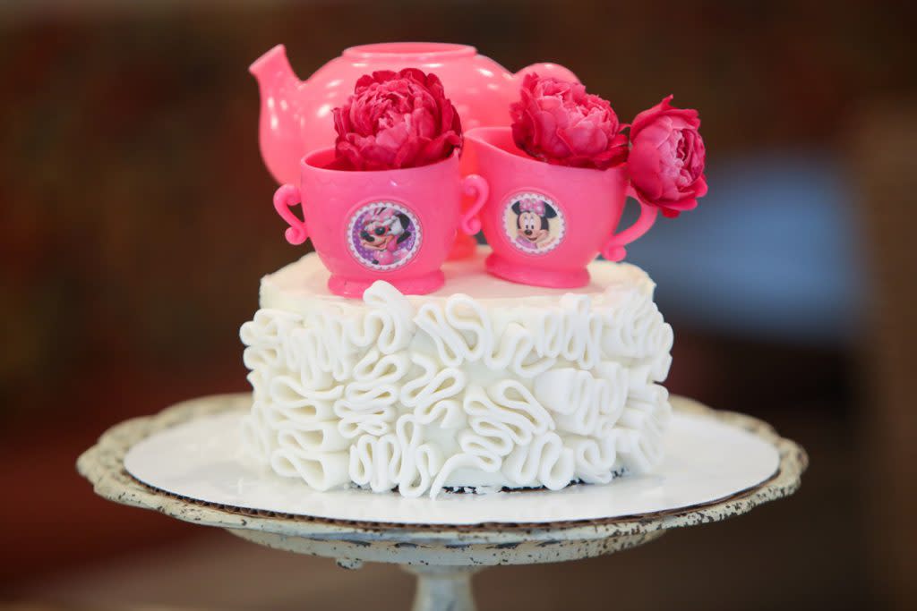 A cake is decorated with fondant bows, a Minnie Mouse tea set and roses 