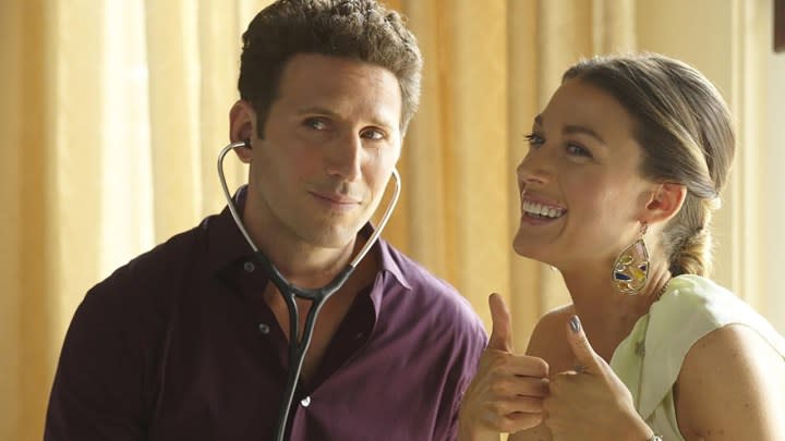 A man wearing a stethoscope stands beside a woman smiling and giving two thumbs up in a scene from Royal Pains.