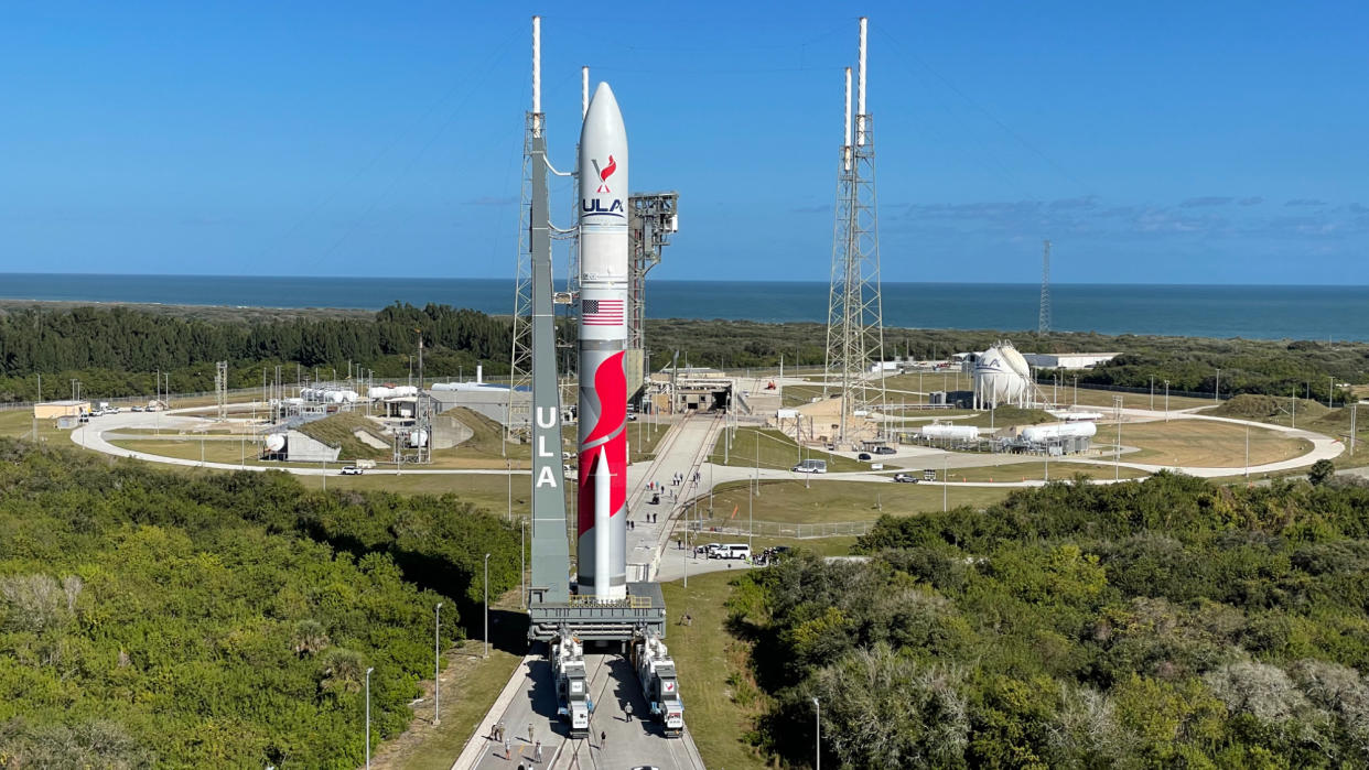  A red and white rocket rolls toward a launch pad, with trees and the ocean in the distance. 