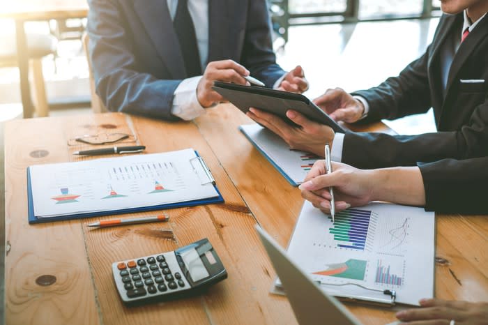 Two men in businesswear sit at a table reviewing financial documents.