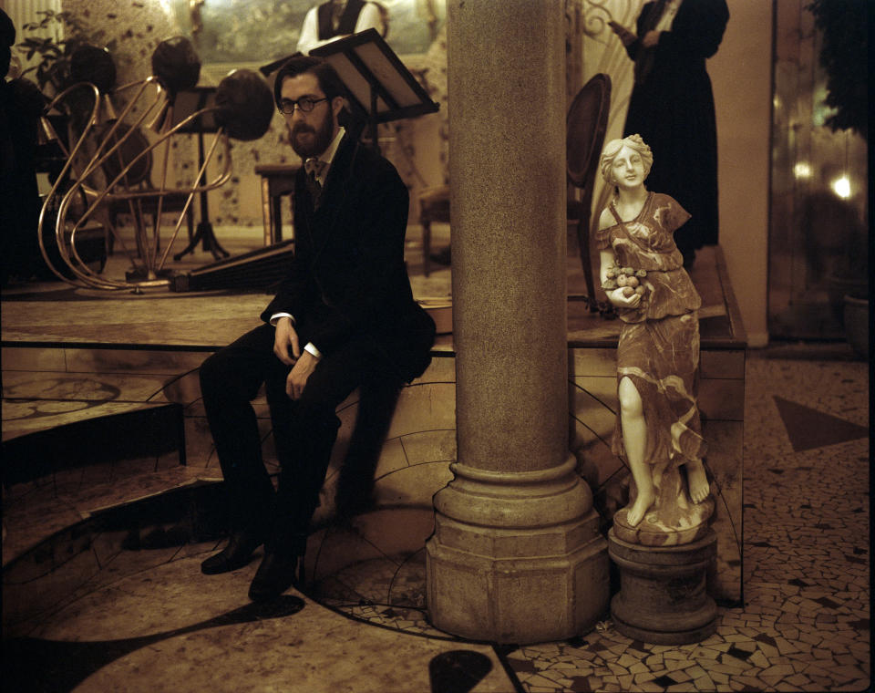 Composer Jerskin Fendrix in a suit and bowtie sitting in front of a bandstand on the set of "Poor Things." 