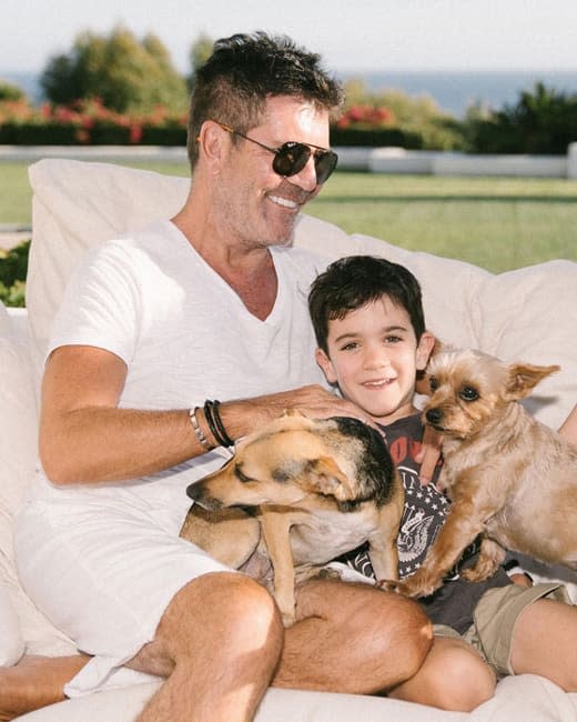 simon and a tiny boy who looks exactly like him sit on a cream sofa with two dogs and a large lawn can be seen behind them