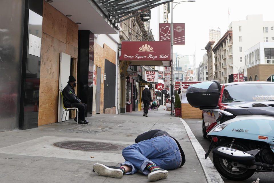 A man lies on the street as a security guard sits in front of the entrance to the Hotel Diva in San Francisco, Saturday, Nov. 21, 2020. Some counties in California are pushing ahead with plans to wind down a program that's housed homeless people in hotel rooms amid the pandemic, despite an emergency cash infusion from the state aimed at preventing those same residents from returning to the streets in cold, rainy weather as the virus surges. (AP Photo/Jeff Chiu)