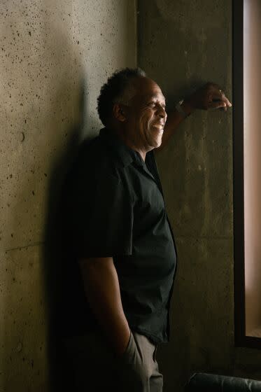 Henry Taylor, wearing a black shirt and leaning on a concrete wall, is illuminate by a small beam of sunlight