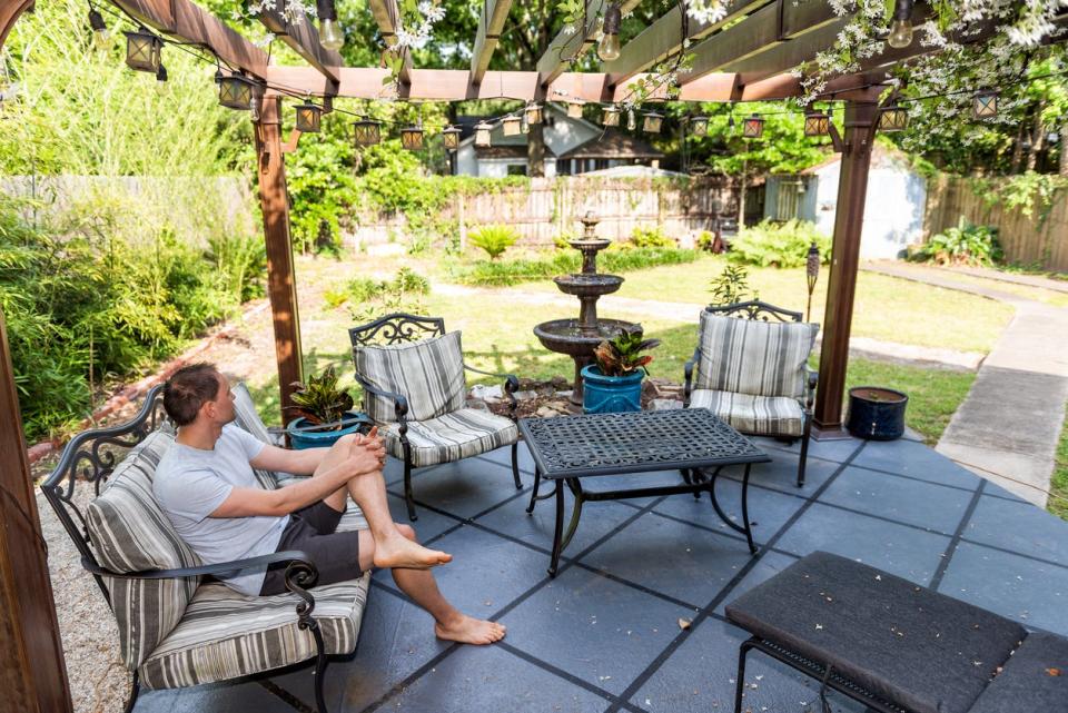 Man sitting under backyard pergola with dark patio furniture and fountain. 