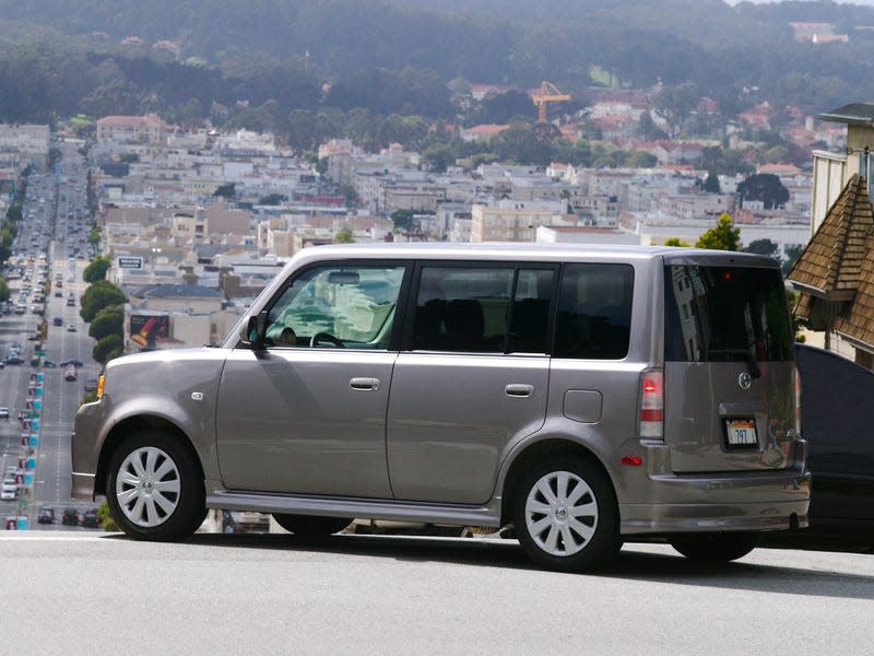 A first-generation gray Scion xB seen from the rear quarter, overlooking a city street from the top of a hill.