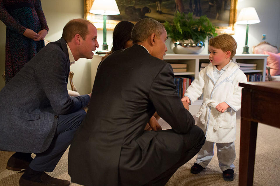 Prince William kneels beside President Obama who is chatting with a young boy in a white robe, in an elegant room