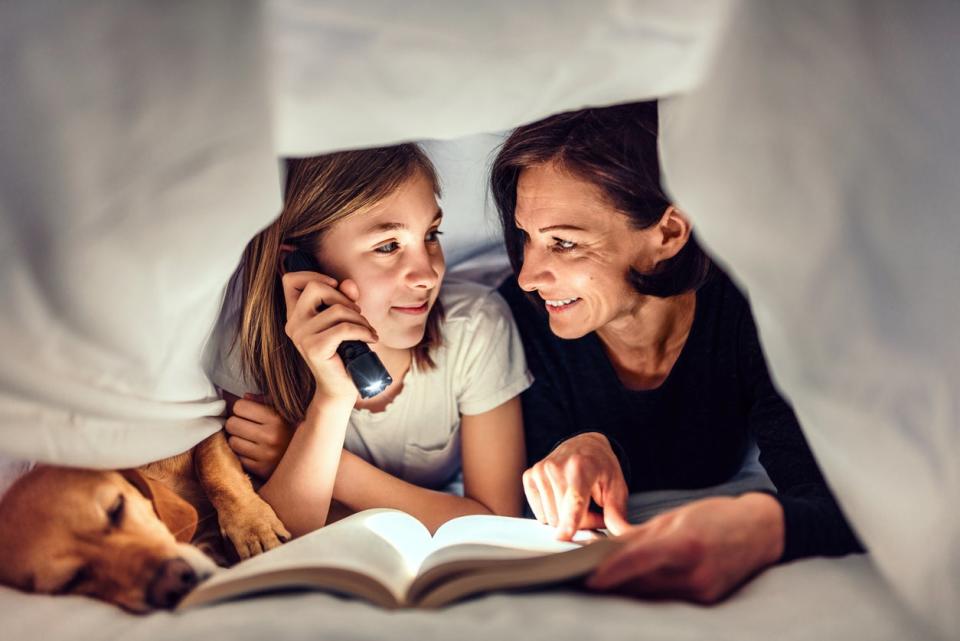 Mother, daughter and dog laying on the bed under blanket holding flashlight and reading book late at night