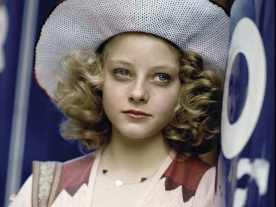 A young Jodie in wide brimmed hat, curly hair, and makeup on leaning against a wall.