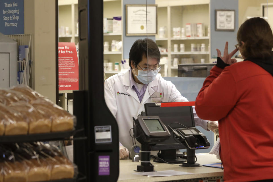 A pharmacist, left, assists a customer while working behind a plastic shield at a grocery store, Thursday, March 26, 2020, in Quincy, Mass. Grocery stores across the U.S. are installing protective plastic shields at checkouts to help keep cashiers and shoppers from infecting each other with the coronavirus.  (AP Photo/Steven Senne)