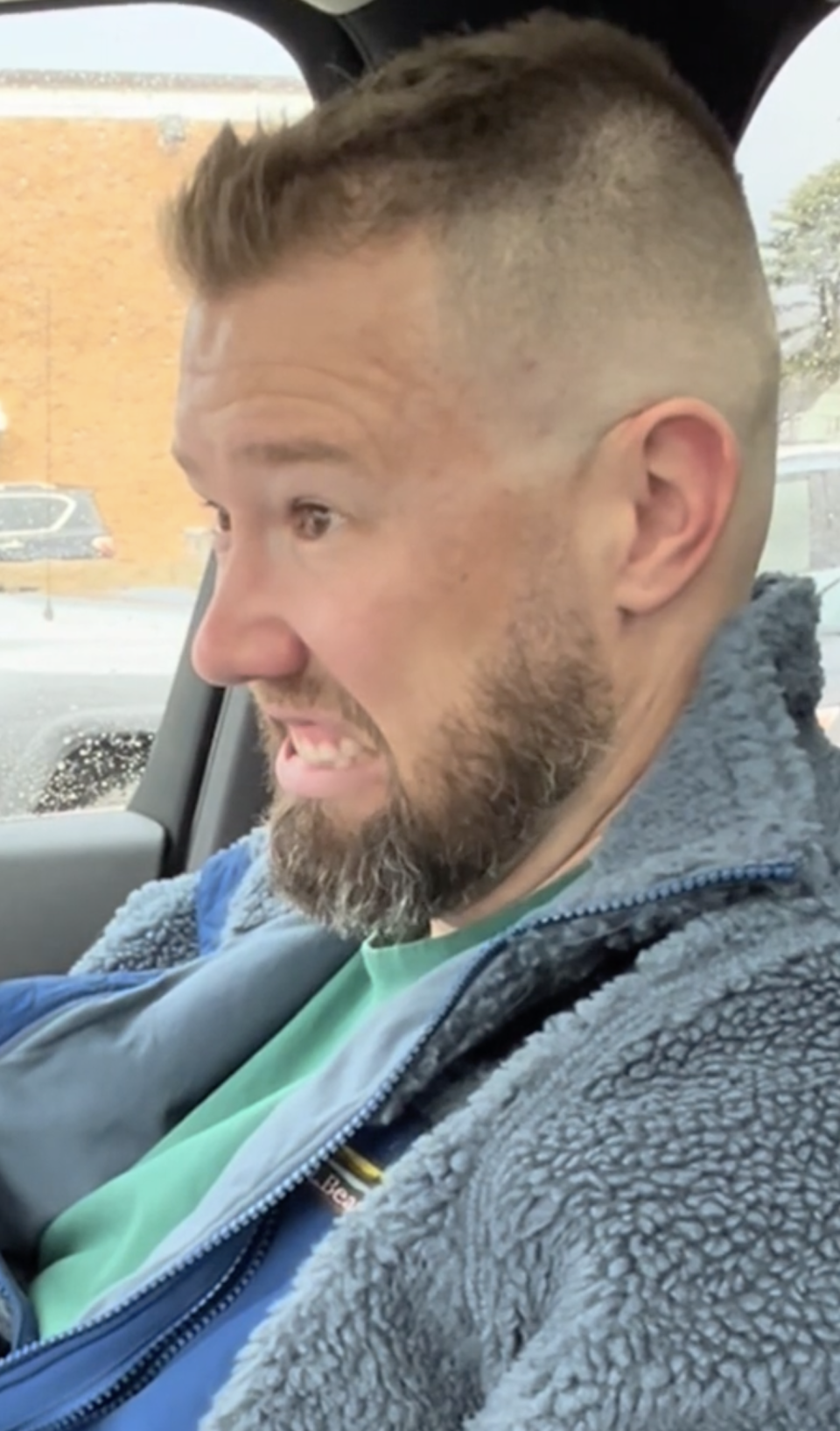 Man with short hair and beard makes a grimacing face while sitting in a car, wearing a zip-up jacket