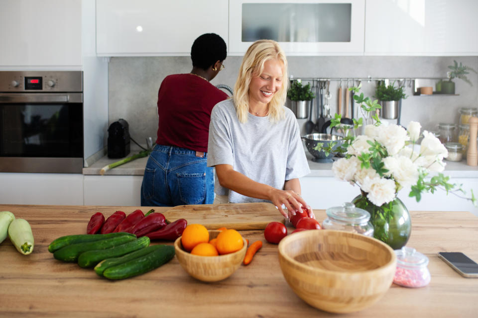 Two women in a kitchen; one is cutting vegetables and the other is washing dishes. A variety of fruits and vegetables are on the countertop