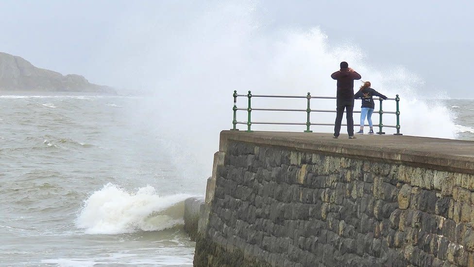 Two people watch the stormy seas at the end of a pier in Criccieth, Wales