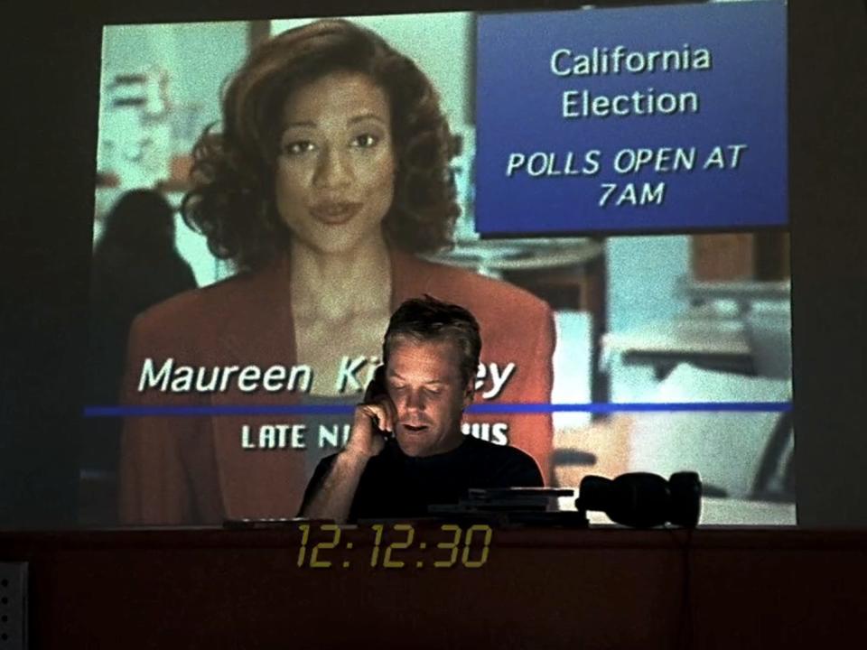 kiefer sutherland as jack bauer on 24, standing in front of a newscast projected on a wall, talking on the phone