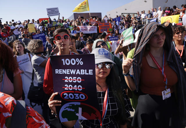 A young protester in a large group holds a poster that reads: 100% Renewable, with a red circle around the year 2030 (and 2040 and 2050 under erasure). 
