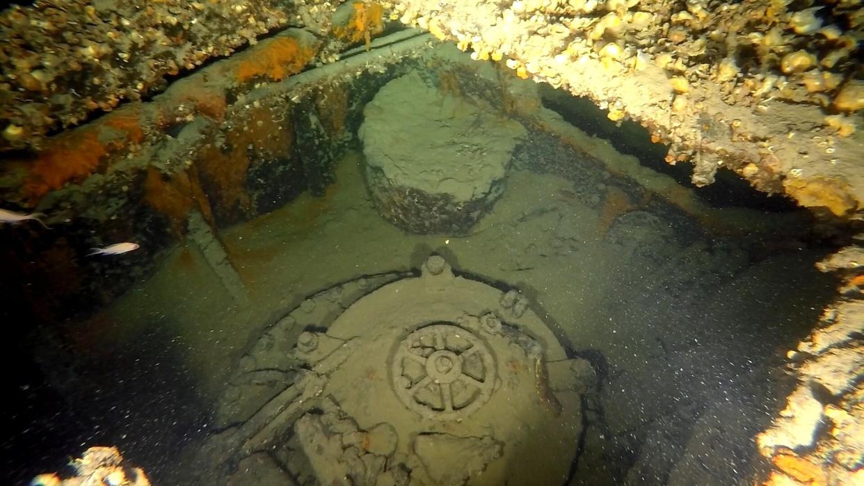  We see an underwater wreck of a submarine. The hatch is closed and sea life is growing on the metal of the sub. 