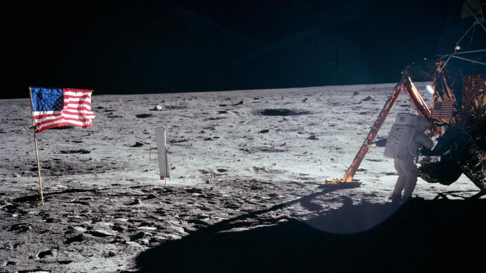     An astronaut in a spacesuit with a gold face shield walks across the gray, dusty surface of the moon. 