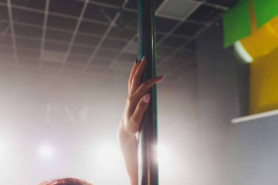 A close-up image of a person's hand grasping a pole, suggesting a sensual or intimate setting. The background is blurred and the person's face is not visible